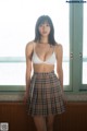 A woman in a white bra top and plaid skirt posing by a window.